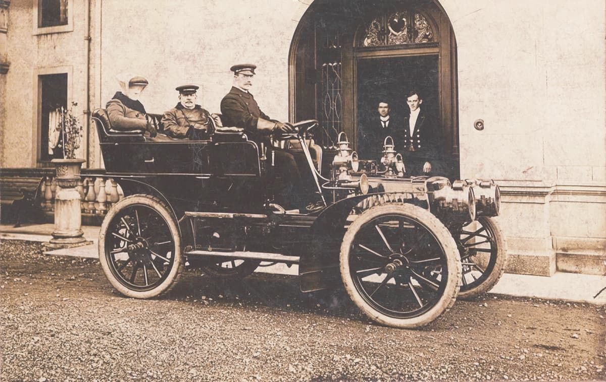 Black and white photograph of Sir Charles and Lady Cayzer, with driver, in their car at Gartmore . Two staff stood in entrance to house.